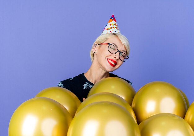 Sonriente joven rubia fiestera con gafas y gorra de cumpleaños de pie detrás de globos con los ojos cerrados aislados sobre fondo púrpura