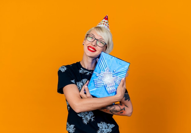 Sonriente joven rubia fiestera con gafas y gorro de cumpleaños con caja de regalo mirando a cámara aislada sobre fondo naranja con espacio de copia