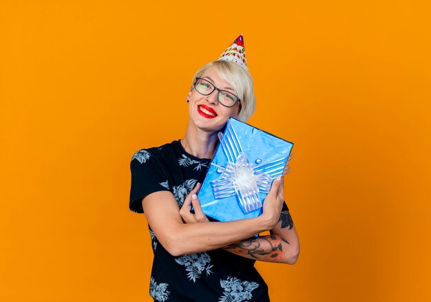 Sonriente joven rubia fiestera con gafas y gorro de cumpleaños con caja de regalo mirando a cámara aislada sobre fondo naranja con espacio de copia