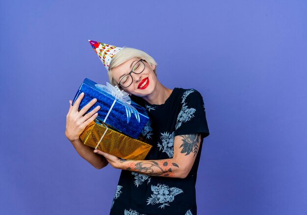 Sonriente joven rubia fiestera con gafas y gorro de cumpleaños sosteniendo cajas de regalo poniendo la cabeza sobre ellos con los ojos cerrados aislados sobre fondo púrpura con espacio de copia