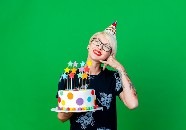 Sonriente joven rubia fiestera con gafas y gorro de cumpleaños sosteniendo pastel de cumpleaños con estrellas mirando al lado haciendo gesto de llamada aislado sobre fondo verde con espacio de copia