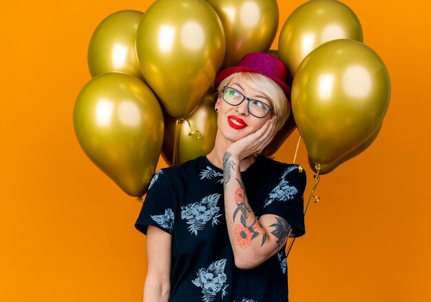 Sonriente joven rubia fiestera con gorro de fiesta y gafas de pie delante de globos tocando la cara mirando al lado aislado sobre fondo naranja