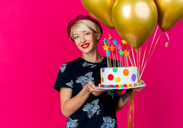 Sonriente joven rubia fiestera con gorro de fiesta sosteniendo globos y pastel de cumpleaños con estrellas mirando a cámara aislada sobre fondo carmesí