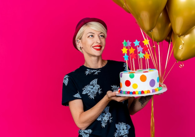 Foto gratuita sonriente joven rubia fiestera con gorro de fiesta sosteniendo globos y pastel de cumpleaños con estrellas mirando globos aislados sobre fondo carmesí con espacio de copia