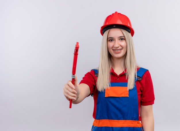 Sonriente joven rubia ingeniero constructor chica en uniforme en aparatos dentales estirando llave de tubo en espacio en blanco aislado con espacio de copia