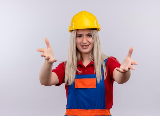 Sonriente joven rubia ingeniero constructor chica en uniforme en aparatos dentales estirando las manos en el espacio en blanco aislado