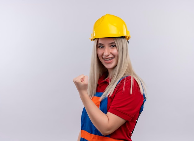 Sonriente joven rubia ingeniero constructor chica en uniforme en aparatos dentales levantando el puño en un espacio en blanco aislado con espacio de copia