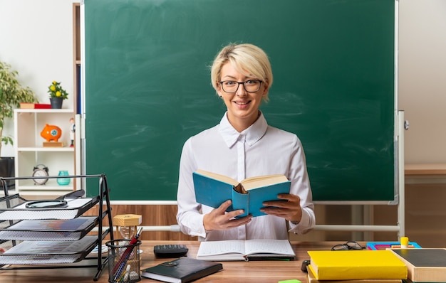 Sonriente joven rubia maestra con gafas sentados frente al escritorio con útiles escolares en el aula sosteniendo el libro abierto mirando al frente