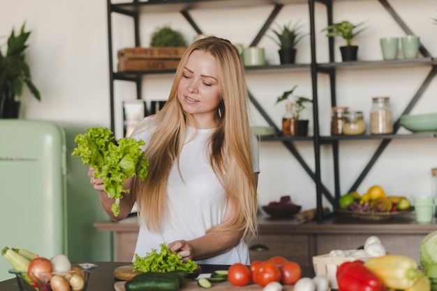 Sonriente joven rubia de pie en la cocina de clasificación de lechuga