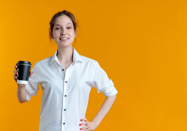 Sonriente joven rubia rusa sostiene la taza de café aislada en el espacio naranja con espacio de copia