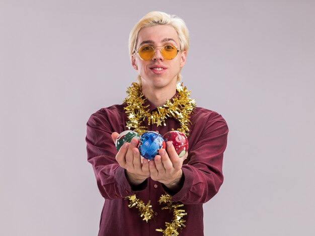 Sonriente joven rubio con gafas con guirnalda de oropel alrededor del cuello estirando adornos navideños hacia la cámara mirando a cámara aislada sobre fondo blanco con espacio de copia