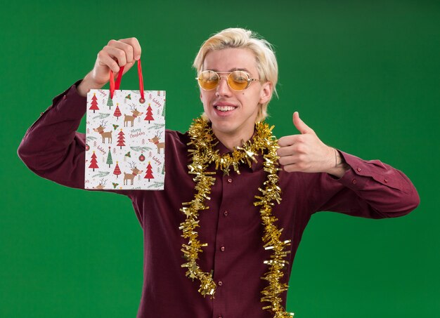 Sonriente joven rubio con gafas con guirnalda de oropel alrededor del cuello sosteniendo una bolsa de regalo de Navidad mostrando el pulgar hacia arriba aislado en la pared verde