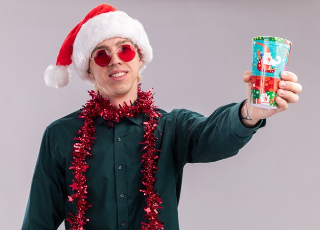 Sonriente joven rubio con gorro de Papá Noel y gafas con guirnalda de oropel alrededor del cuello estirando la taza plástica de Navidad hacia la cámara mirando a cámara aislada sobre fondo blanco