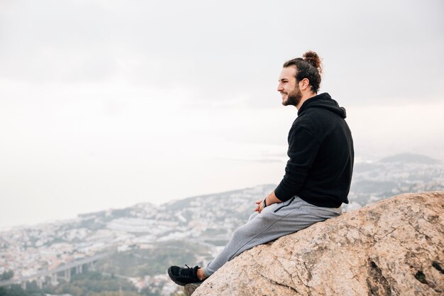 Sonriente joven sentado en el pico de la montaña mirando el paisaje urbano