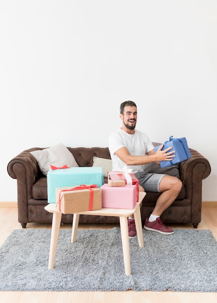 Sonriente joven sentado en el sofá con caja de regalo azul en casa