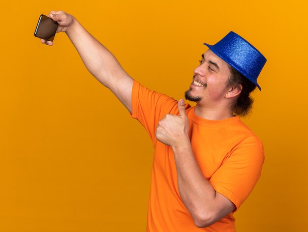 Sonriente joven con sombrero de fiesta tomar un selfie mostrando el pulgar hacia arriba aislado en la pared naranja