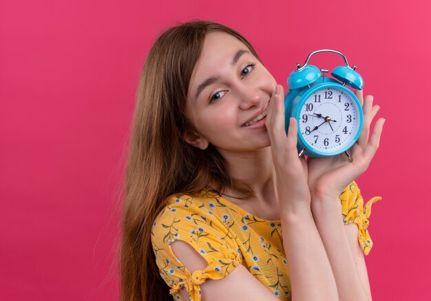 Sonriente joven sosteniendo el despertador en la pared rosa aislada