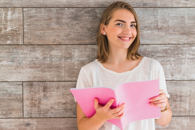 Sonriente joven sosteniendo libro rosa contra la pared de madera