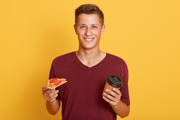 Sonriente joven sosteniendo llevar café y rebanada de pizza sabrosa ,, merienda, vistiendo camiseta marrón