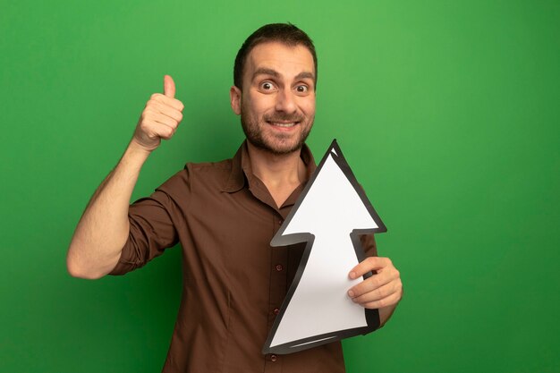 Sonriente joven sosteniendo la marca de la flecha que apunta hacia arriba mirando al frente mostrando el pulgar hacia arriba aislado en la pared verde