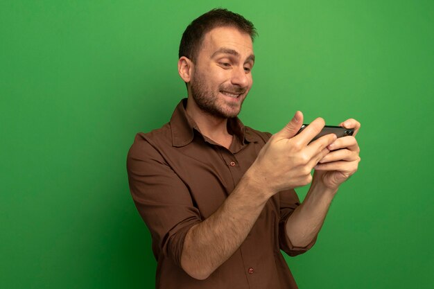 Sonriente joven sosteniendo y mirando el teléfono móvil aislado en la pared verde