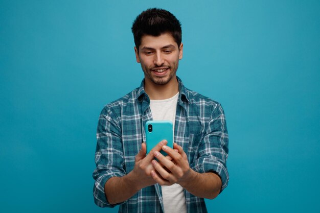Sonriente joven sosteniendo y mirando el teléfono móvil aislado sobre fondo azul.
