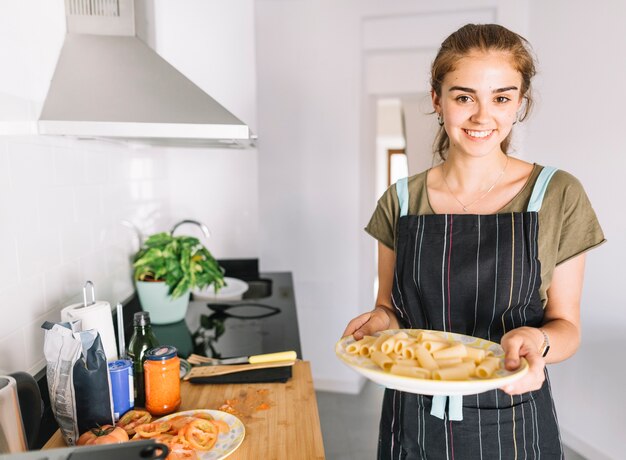 Sonriente joven sosteniendo el plato de pasta rigatoni crudo en la cocina