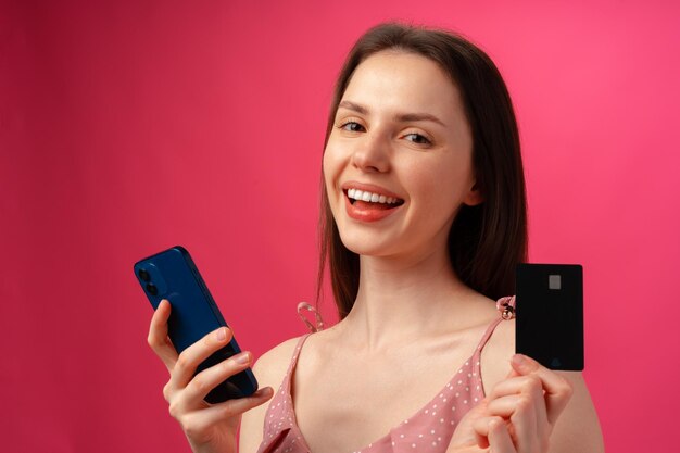 Sonriente joven sosteniendo smartphone y tarjeta de crédito contra fondo rosa