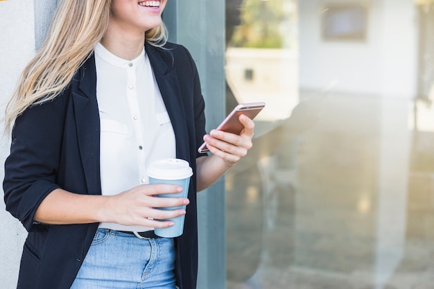 Sonriente joven sosteniendo la taza de café de papel y teléfono móvil