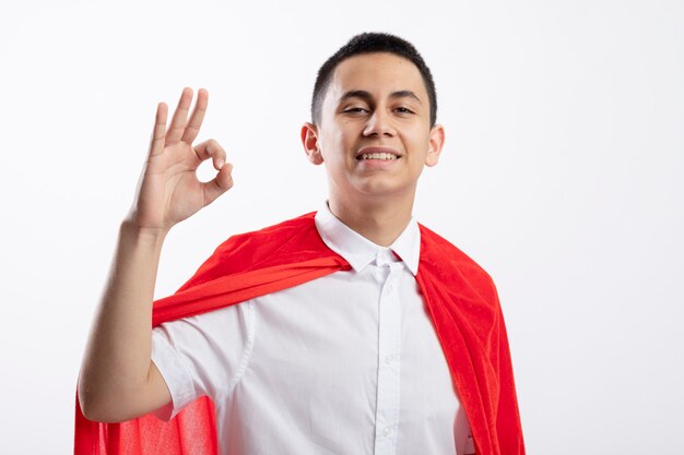 Sonriente joven superhéroe en capa roja mirando a cámara haciendo bien signo aislado sobre fondo blanco con espacio de copia