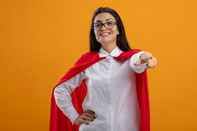 Foto gratuita sonriente joven superhéroe caucásica con gafas mirando y apuntando a la cámara manteniendo la mano en la cintura aislada sobre fondo naranja con espacio de copia