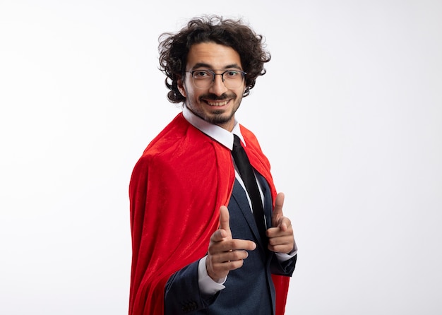 Sonriente joven superhéroe caucásico en gafas ópticas con traje con capa roja se encuentra de lado apuntando a la cámara con las dos manos aisladas sobre fondo blanco con espacio de copia