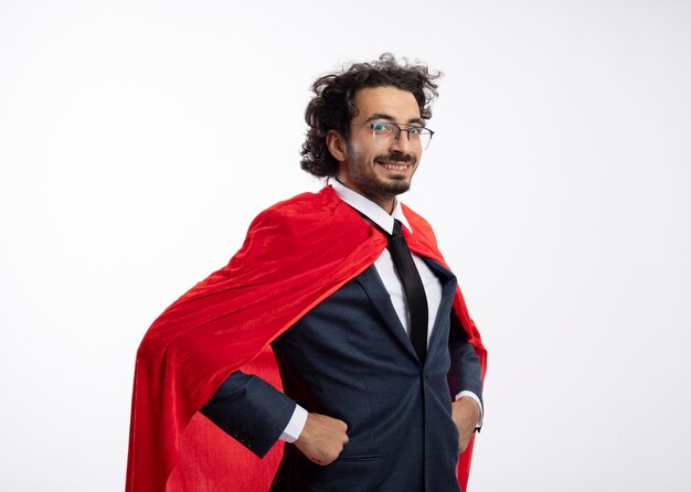 Sonriente joven superhéroe caucásico en gafas ópticas con traje con capa roja se encuentra de lado poniendo las manos en la cintura aislada sobre fondo blanco con espacio de copia