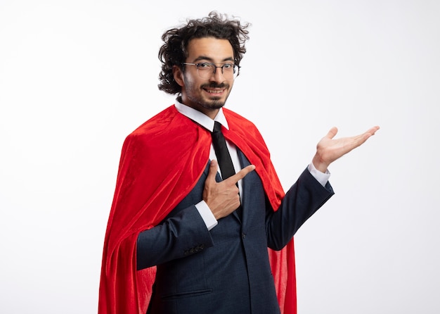 Sonriente joven superhéroe caucásico en gafas ópticas con traje con manto rojo apunta en la mano vacía