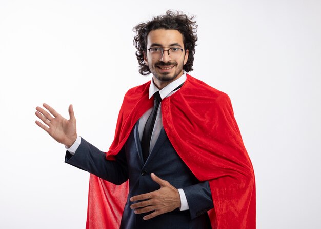Sonriente joven superhéroe caucásico en gafas ópticas con traje con manto rojo se encuentra de lado con la mano levantada