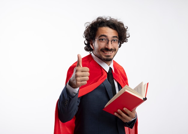 Sonriente joven superhéroe caucásico en gafas ópticas con traje con manto rojo sostiene libro y pulgar hacia arriba