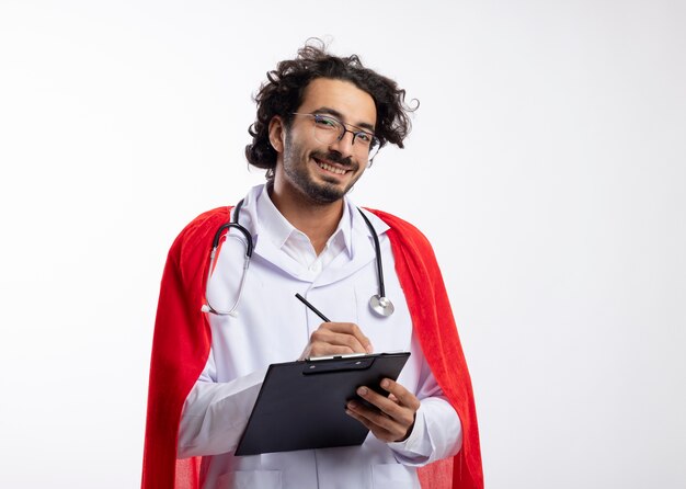 Sonriente joven superhéroe caucásico con gafas ópticas vistiendo uniforme médico con manto rojo y con estetoscopio alrededor del cuello sostiene lápiz y portapapeles