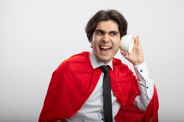 Sonriente joven superhéroe chico mirando al lado con corbata mostrando escuchar gesto con taza aislado sobre fondo blanco.