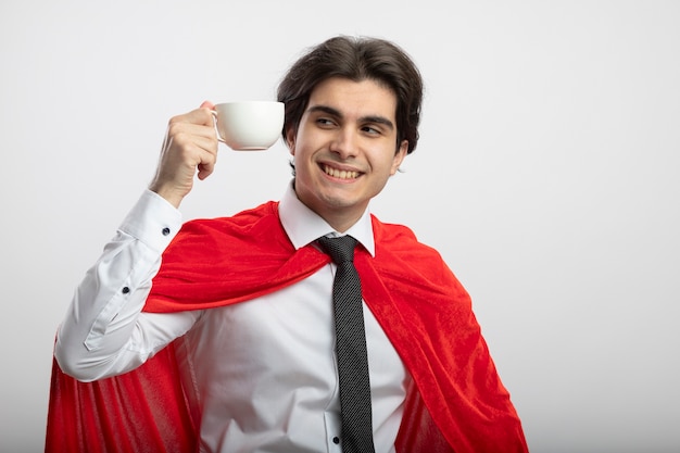 Sonriente joven superhéroe chico vistiendo corbata sosteniendo y mirando una taza de café aislado sobre fondo blanco.