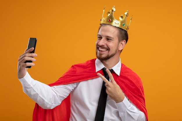 Foto gratuita sonriente joven superhéroe con corbata y corona mostrando gesto de paz tomar un selfie aislado en naranja