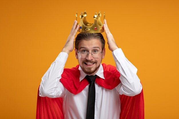 Sonriente joven superhéroe con corbata y gafas poniendo corona en la cabeza aislada en naranja