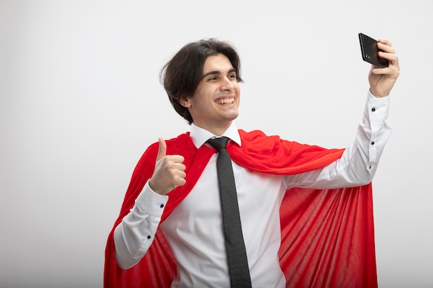 Sonriente joven superhéroe con corbata tomar un selfie mostrando el pulgar hacia arriba aislado sobre fondo blanco.