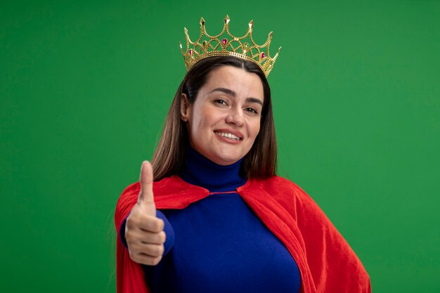 Sonriente joven superhéroe con corona mostrando el pulgar hacia arriba aislado sobre fondo verde