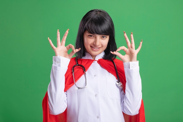 Sonriente joven superhéroe con estetoscopio con bata médica y manto mostrando gestos bien aislados en verde