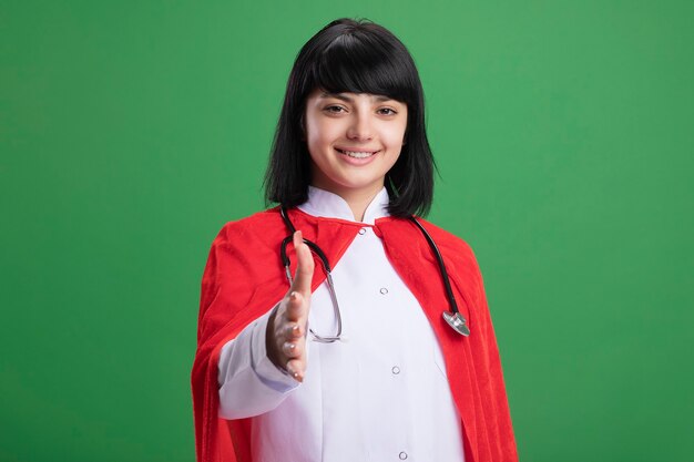 Sonriente joven superhéroe con estetoscopio con bata médica y manto saludo aislado en verde