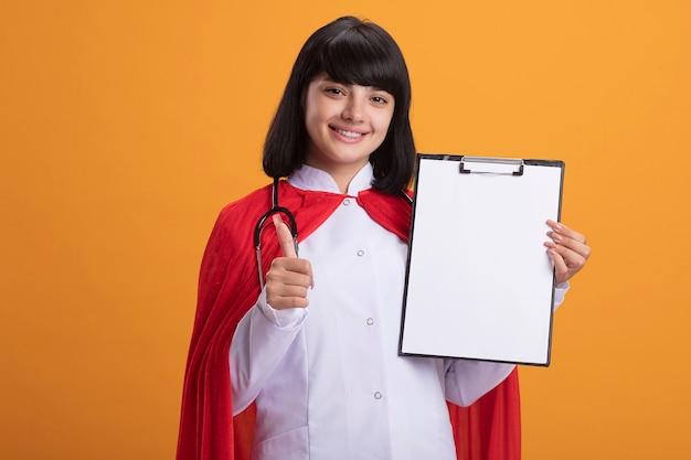 Sonriente joven superhéroe con estetoscopio con bata médica y manto sosteniendo el portapapeles mostrando el pulgar hacia arriba aislado en la pared naranja