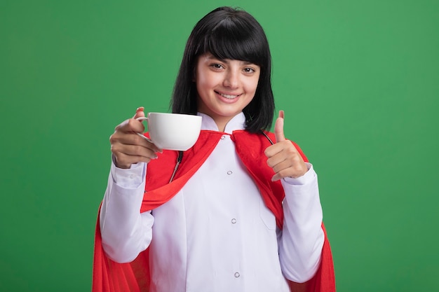 Sonriente joven superhéroe con estetoscopio con bata médica y manto sosteniendo una taza de té mostrando el pulgar hacia arriba aislado en verde