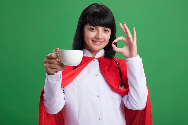 Sonriente joven superhéroe con estetoscopio con bata médica y manto sosteniendo una taza de té que muestra un gesto bien aislado en la pared verde
