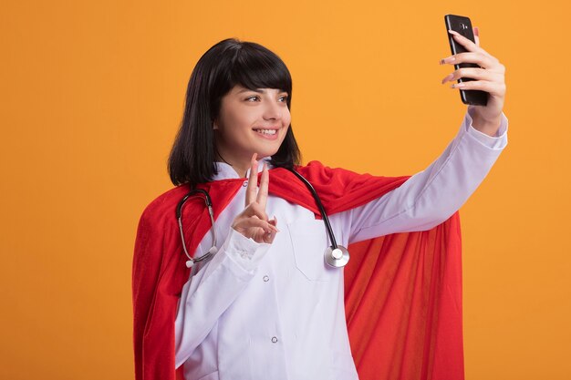 Sonriente joven superhéroe con estetoscopio con bata médica y manto tomar un selfie mostrando gesto de paz