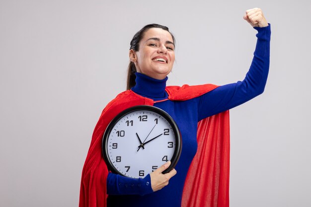 Sonriente joven superhéroe sosteniendo un reloj de pared que muestra un gesto fuerte aislado sobre fondo blanco.